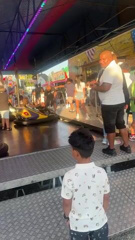 The video shows a bumper car ride at an outdoor amusement park. There are several people on and around the ride. In the foreground, a young boy wearing a white shirt with a pattern of palm trees and surfboards is standing, looking towards the bumper cars. To his right, a man with a shaved head and a white t-shirt and dark shorts is standing and talking to a woman in a pink dress. In the background, bumper cars are moving, and several other people are visible on the ride and in the surrounding area. The ride is decorated with colorful lights and banners. The overall atmosphere appears to be lively and fun.