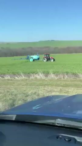 The view from inside a blue vehicle looks out over a green field. A red tractor pulls a turquoise sprayer across the field, leaving a trail of mist behind it. The sky is clear and blue. Behind the field, there are trees. The day appears bright and sunny.