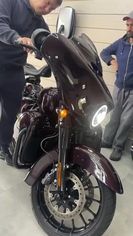 The video shows a close-up view of a dark-colored motorcycle, likely a Harley-Davidson, with its headlight illuminated. Two men are present, one on the left side of the motorcycle, appears to be adjusting something on the handlebars. The other man is standing on the right side of the motorcycle, with his hands on his hips. The motorcycle is parked indoors, possibly in a showroom or garage, with a light-colored, wood-paneled wall in the background. The motorcycle has a large fairing, a prominent headlight, and chrome accents. The video pans slightly to the left, then to the right, focusing on the front of the motorcycle and its details.