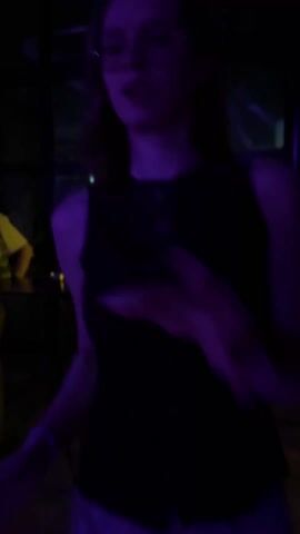 A woman wearing glasses and a sleeveless animal-print shirt is seen dancing in a dimly lit club. The lighting is primarily purple and the dance floor is crowded. There are high cocktail tables in the background, and glimpses of a city skyline can be seen through the windows. The camera focuses on the woman as she moves to the music. Another person’s hand briefly enters the frame in the foreground at the start and end of the video.