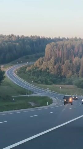 The video shows a highway curving through a forested landscape during daylight. Several cars are visible on the highway. The sun appears to be low in the sky, casting long shadows. The road is paved and has lane markings. The surrounding vegetation is lush and green, consisting of trees and grass. The sky is a pale blue, suggesting either early morning or late afternoon. There are no people visible in the video, only vehicles on the road. The overall impression is one of travel and open space.