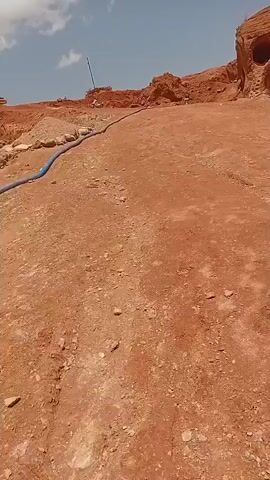 The video pans across a dirt path leading towards a quarry. The ground is red dirt with scattered rocks and debris. A blue hose runs along the left side of the path. In the background, there are piles of dirt and rock, and a large rock formation with several circular openings. A person can be seen standing on top of the rock formation. The sky is blue with some white clouds.