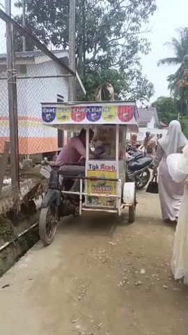 A three-wheeled cart, decorated with colorful banners depicting ice cream and advertising "Sehat Tanpa Pemanis Buatan" (Healthy Without Artificial Sweeteners) and "Tgk Aceh," is parked on a dirt path. A man in a pink shirt sits on the cart, facing a display of ice cream products. To the right, three individuals wearing hijabs are standing near a motorcycle, appearing to be customers. The cart has a white structure with a metal frame and wheels. The background shows a fence, some buildings, and trees, suggesting an outdoor setting. The video appears to be a short clip, possibly a recording of a street vendor.