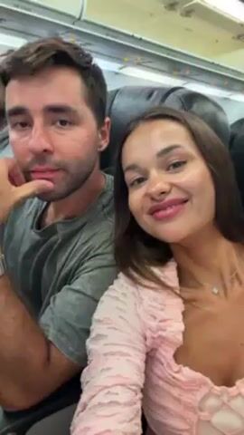 A man and a woman are in a plane, sitting in seats. They both smile for the camera. The man makes a hand gesture. The woman leans in to say something to the man, and then the camera is covered with a pink article of clothing.