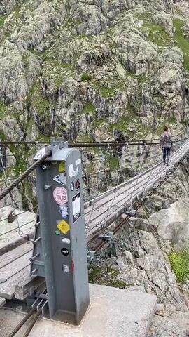 A wide shot of a person walking across a long suspension bridge, extending across a rocky mountain valley. The person, dressed in a grey top and dark pants, is moving from left to right across the bridge. The bridge features wooden planks and metal cables, secured to a large metal pillar in the foreground adorned with stickers. The valley is characterized by steep, grey, rocky slopes covered with patches of green vegetation, and a small waterfall is visible in the background, originating from snow-capped peaks. The sky is overcast.