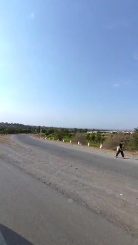 The video shows a view from a moving vehicle traveling on a road. The road is paved and appears to be in a rural or semi-rural area. On the right side of the road, there is a stretch of dry, uncultivated land with sparse vegetation and a few scattered trees. Further in the distance, there are fields that appear greener, possibly cultivated land or grass. A line of white posts or markers runs along the edge of the road, bordering the greener area. On the left side of the road, there are more trees and vegetation, with some slopes and variations in terrain. The sky is clear and blue, suggesting fair weather. The overall impression is of a journey through a landscape with natural elements and infrastructure like roads and markers.