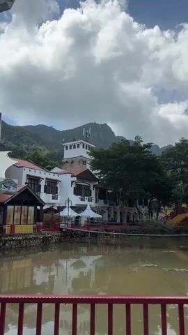 The video shows a scenic view of a body of water in the foreground with a red railing. In the middle ground, there are buildings with white walls and red roofs, surrounded by lush greenery and palm trees. A distinctive tower with a cross-shaped structure on top is visible behind the buildings. Several tents and a red bridge add to the landscape. In the background, mountains rise majestically under a sky filled with dramatic clouds. The water reflects the surrounding scenery, creating a peaceful and picturesque atmosphere. The camera pans slightly to the right, revealing more of the park-like setting with various trees and architectural elements.