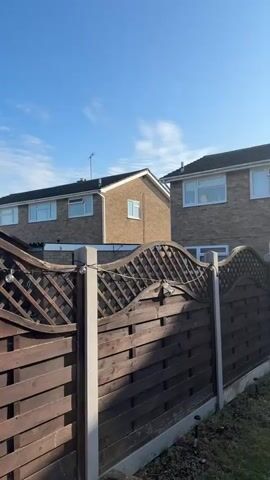 The media file presents a panoramic view of a residential backyard in Colchester, United Kingdom, on a bright, sunny day. The scene begins with a close-up of a dark-stained wooden fence, supported by grey concrete posts, featuring a decorative wavy lattice top section. The ground immediately adjacent to the fence appears to be bare earth or disturbed soil.

As the camera pans from left to right, a neatly maintained lawn comes into view, traversed by a winding path made of irregular-shaped grey stepping stones. Beyond the lawn and patio, the rear of a two-story brick house is visible. This house features white-framed windows and a cream-colored single-story extension with white double French doors. A tall, slender, neatly trimmed hedge-like plant stands next to the doors, and a green garden hose reel is partially visible nearby. An old-fashioned television antenna is mounted on the roof of this house.

The pan continues to reveal a second, similar two-story brick house to the right, also with white windows and an attached single-story section. The sun is high and bright in the sky, creating a strong lens flare in the upper right portion of the frame and casting distinct, long shadows across the grass, suggesting either morning or late afternoon. The sky is clear blue with only a few faint wisps of white cloud. No people or visible text are present in the scene. The overall impression is one of a quiet, well-maintained suburban garden under clear, sunny weather.