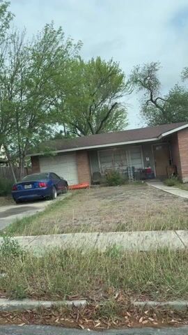 The video shows a drive through a neighborhood on a cloudy day. The camera is inside a moving car. The houses are single-story and have front yards with varying levels of landscaping. Some have cars parked in the driveways or carports. There is a red car next to the car the camera is in. The street is paved and has concrete curbs. Some trees are scattered throughout the scene. There is a hotel in the distance.