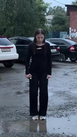 A young woman with long dark hair is jumping in a puddle. She is wearing a black long-sleeved shirt and black pants. She is also wearing white sneakers. She is holding a yellow object in her right hand and a black purse in her left hand. The puddle is on a gravelly surface. There are cars parked in the background. The weather appears to be overcast. The woman is in motion, with her legs bent and her body angled as she jumps.