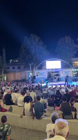 A nighttime outdoor concert is taking place on a stage decorated with banners. A band is performing on stage, with musicians playing instruments and singing. The stage is lit with colorful lights. A large audience is seated in front of the stage, watching the performance. The audience members are of various ages and are dressed casually. The environment appears to be a public space, possibly a plaza or park, with trees and buildings visible in the background. The sky is dark, indicating it is night.