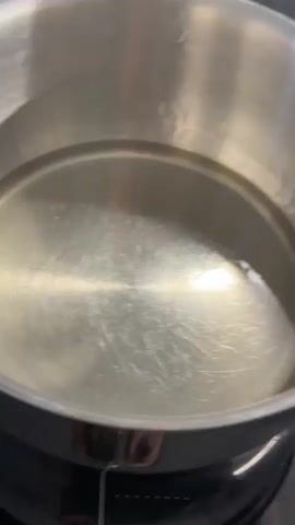 A hand with manicured nails is adding several ridged potato slices to a pot of boiling water. The potato slices are thin and round, with a ribbed texture. They are being added one by one and are beginning to float in the water. The pot is silver and sits on a black stovetop. The water is bubbling vigorously, indicating it is hot. Some of the potato slices appear slightly translucent.