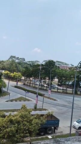 THE VIDEO SHOWS A VIEW OF A SEMI-URBAN INTERSECTION ON A BRIGHT DAY. THE SKY IS MOSTLY CLEAR WITH A FEW CLOUDS. TREES LINE THE SIDES OF THE ROAD AND AROUND THE INTERSECTION. A FEW CARS ARE VISIBLE ON THE ROAD, INCLUDING A WHITE SUV AND A WHITE SEDAN. A BLUE DUMP TRUCK IS PARKED NEAR THE BOTTOM OF THE FRAME. MALAYSIAN FLAGS ARE DISPLAYED ON POLES ALONG THE ROADWAY. THE INTERSECTION HAS MARKED LANES AND ROAD MARKINGS. THE OVERALL SCENE SUGGESTS A TYPICAL DAYTIME VIEW IN A DEVELOPED AREA.