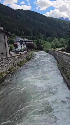 A wide shot captures a fast-flowing river, its water appearing white and turbulent in sections, as it carves a path through a valley. The river is contained by sturdy stone walls on both banks. On the left bank, several buildings with traditional architecture, including a prominent white house with a dark roof and red accents, are nestled amidst greenery. Parked cars are visible near these structures. The opposite bank also features some trees and what appears to be a road or path.

In the background, lush green mountains covered in dense forest rise steeply under a bright blue sky dotted with fluffy white clouds. The overall impression is one of a picturesque natural landscape with a charming village integrated into it. The sun is shining brightly, suggesting a clear day, likely late morning or afternoon. There is no visible text in the video. The location is Ponte di Legno, Italy.