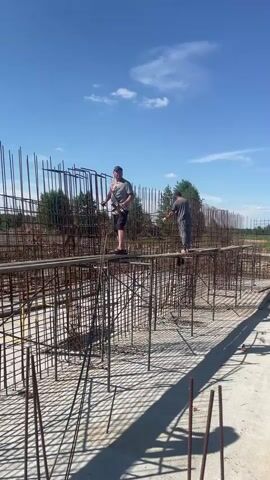 The video shows two men working on a construction site. They are standing on wooden planks, surrounded by a grid of rebar. The rebar is vertical and horizontal, forming a cage-like structure. The men appear to be tying the rebar together with wire. The sky is blue with some clouds, and there are trees in the background. The ground is covered in dirt and gravel. In the later part of the video, a construction vehicle is visible in the distance. There is also a pool of murky water on the ground. The overall scene depicts an active construction project.