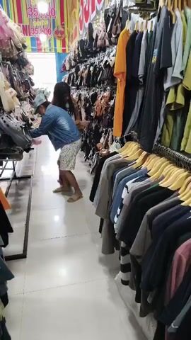 The video shows two people in a store aisle filled with clothing and accessories. On the left, shelves are packed with what appear to be bags and shoes, with a price tag displaying "35,000". A person wearing a blue denim jacket, a light-colored cap, and patterned shorts is bent over, examining items on a low shelf. To their right and slightly behind, another person with long dark hair, wearing a brown jacket and a white t-shirt, stands and observes. The right side of the aisle features racks of hanging clothes, predominantly t-shirts and long-sleeved shirts in various colors like orange, black, blue, and gray. The store appears brightly lit with a colorful striped background visible at the far end.