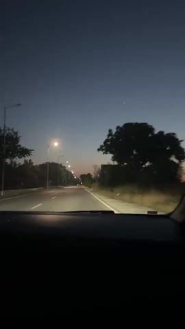 The video is a first-person perspective from inside a car traveling on a road at dusk or night. The road is illuminated by streetlights and the headlights of the car. The sky is a deep blue, transitioning to a faint orange hue near the horizon, suggesting either sunrise or sunset. There are trees and vegetation on both sides of the road, and a few distant lights indicate a populated area or passing vehicles. The dashboard of the car is visible in the foreground.