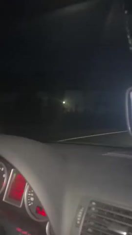 The video shows a night-time view from inside a vehicle, likely a car. The camera is pointed forward, through the windshield. The interior of the car is dark, but the dashboard lights are visible, displaying red illumination. Outside the car, the scene is dark with a few distant lights, including what appears to be a street lamp and some faint headlights. The road ahead is visible, with a white line marking the lane. The overall impression is of driving on a dark road at night.