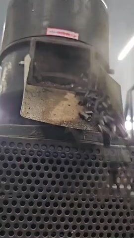 A close-up, low-angle shot shows a machine with a metal hopper filled with dark, cylindrical pellets that appear to be burning or smoking. The pellets are being fed from the hopper into a lower section of the machine, which has a perforated metal grate. Smoke is visible rising from the top of the machine. The pellets are small, roughly the size of a finger, and are uniform in shape. The machine itself is dark gray and appears to be industrial in nature. The background is blurred, but suggests an indoor environment with artificial lighting. The video is short, lasting only a few seconds, and captures the continuous feeding and processing of the pellets. There are no people visible in the frame.