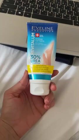 A hand is holding a tube of Eveline Cosmetics Revitalum 30% Urea Complex cream mask against a white, textured surface, possibly a bed or blanket. In the background, a laptop keyboard is visible. The cream mask tube is blue and white with yellow and turquoise accents. It features an image of a foot and text indicating its purpose for intense softness and against calluses. The lighting is bright, suggesting it's daytime or a well-lit indoor setting.
