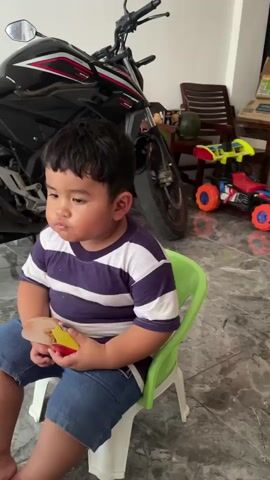 A young boy sits on a green plastic chair holding a red and yellow object, possibly a book or toy. He is wearing a striped t-shirt and denim shorts. Behind him is a black motorcycle. In the background, there are wooden chairs and a toy car. The floor is tiled. The boy appears to be chewing or eating something. The lighting suggests an indoor setting.