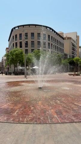 This is a short, vertical video of a modern public square with a water fountain. The fountain is the central subject, spraying water upwards in a wide arc, creating a mist that catches the sunlight. The ground around the fountain is paved with reddish-brown tiles arranged in a circular pattern, with the water pooling and reflecting the light. Beyond the fountain, there is a paved area with some trees and streetlights.

In the background, a large, modern building with a curved facade dominates the scene. The building has many windows and appears to be multi-story. To the left of the building, there are more trees and hints of outdoor seating areas, suggesting a commercial or residential space. A few people are visible in the distance on the left, walking or sitting, and a bicycle rack is also present.

The time of day appears to be daytime, likely late morning or early afternoon, given the bright sunlight and clear blue sky. The weather seems to be warm and sunny. The overall impression is one of a pleasant, open public space designed for relaxation and enjoyment. The architectural style of the building and the paved plaza suggest a contemporary urban setting. There is no visible text in the video. The location is identified as Almería, Spain.