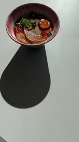 The video shows a bowl of ramen, with red, white, and brown broth, toppings including pork slices, a half-boiled egg, mushrooms, and green scallions. The bowl has a red and white pattern. The bowl is on a white surface, with a shadow cast to its left.  There are background sounds of people talking.