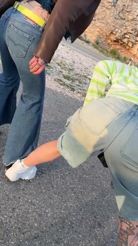 The video features two individuals in an outdoor setting, likely a paved area next to a rocky, mountainous landscape, consistent with Bukovica, Montenegro. The scene is brightly lit by sunlight, suggesting daytime, possibly late afternoon with warm tones.

On the left, a person, likely a woman, is visible from the waist down, wearing blue flared jeans, a brown jacket, and white chunky sneakers. A prominent yellow belt with "DIESEL" written in red letters is cinched at her waist. Rings are visible on her fingers. She stands with a slight forward lean.

On the right, another person, also likely a woman, is captured bending over. She wears light blue denim cargo shorts, a partially visible light green and white striped long-sleeved shirt, and white chunky sneakers. A detailed tattoo adorns her lower leg. A hand rests on her hip or lower back.

The activity appears to be a playful or dynamic pose, possibly part of a dance or performance, accompanied by rhythmic chanting or cheering sounds from off-screen, contributing to an energetic and lively atmosphere.