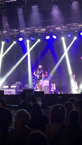 The video shows a concert with a band performing on stage. There are several spotlights illuminating the stage with blue and white light. A lead singer is at the front of the stage playing a guitar and singing. There is a drummer behind him and another band member playing a bass guitar on the right side of the stage. In front of the stage, an audience is visible, silhouetted against the stage lights. The overall atmosphere appears to be energetic and lively.