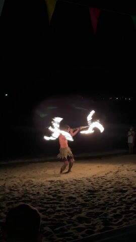 A shirtless man on a beach at night performs a fire dance with two fire sticks. He is wearing patterned shorts and is kneeling on the sand, twirling the sticks in front of him. The sand is light brown and illuminated by the fire, while the background is dark. There are colorful flags hanging above the performance area. Two other people are visible in the background.