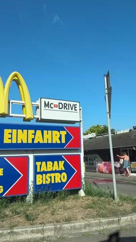 The video begins with a shot of a McDonald's sign in German, indicating "McDrive," "Einfahrt" (entrance), and "Tabak Bistro." The yellow arches of the McDonald's logo are visible. A person wearing a blue top and shorts is seen walking in the background near the McDonald's building. The camera then pans to show a road with a large truck and a smaller orange truck. Finally, the video shows a McDonald's paper bag with "MISSION: WORLD MENU" printed on it. The bag has various stickers representing different countries such as Malaysia, Canada, Japan, and Brazil, along with images of a guitar and a calculator.