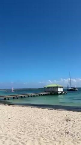 The scene is a sunny, picturesque beach with a clear blue sky. A wooden pier stretches out into the turquoise water, leading to a small, white shack with a green roof. To the left, people are gathered in the water near several sailboats. A larger sailboat is moored to the right of the pier. The beach is sandy and stretches across the bottom of the frame. The overall atmosphere is peaceful and idyllic.