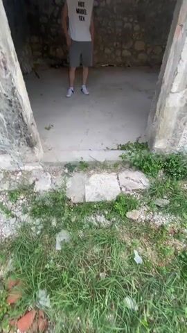 A person with curly brown hair, wearing a white sleeveless t-shirt with "HOT MODEL SEX" printed on it and grey shorts, stands in the middle of a room with stone walls and a wooden ceiling. The room appears to be an unfinished structure, with several windows. The person takes a few steps to the right and looks around the room. The camera then pans around the room, showing the stone walls, windows, and the wooden ceiling. The person is then seen walking towards the back of the room. The video ends with a shot of the room from the back, with the person in the distance.
