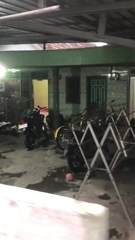 The video shows a residential area in Wunut, Indonesia, at night. The scene is dimly lit, with the primary light sources appearing to be from inside the houses and a few streetlights. The foreground is dominated by several motorcycles parked under a covered area, possibly a carport or the front of a house. Drying racks with laundry are also visible in front of the motorcycles. Beyond the motorcycles, a house with a green facade and a light-colored door is visible. The entrance of the house is illuminated by a warm yellow light, suggesting activity within. Another house, with a darker exterior, is partially visible on the right side of the frame. A car is parked on the right, its headlights faintly illuminated. The ground appears to be concrete, with some puddles of water, indicating recent rain or damp conditions. The overall impression is of a quiet residential street at night, with the typical sights of parked vehicles and a glimpse into domestic life. There is no visible text in the video.