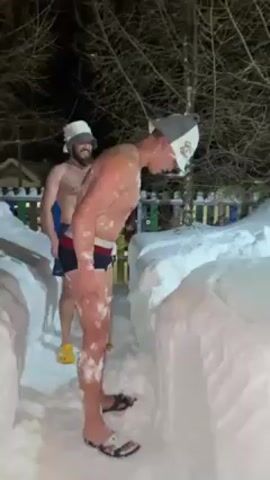 Two shirtless men in swim trunks and felt hats are standing in a snowy area. One man rubs snow on his chest and face. The other man stands behind him and smiles. The snow appears to be banked up on either side of them, forming a sort of pathway. It is nighttime. A fence and a building are visible in the background.