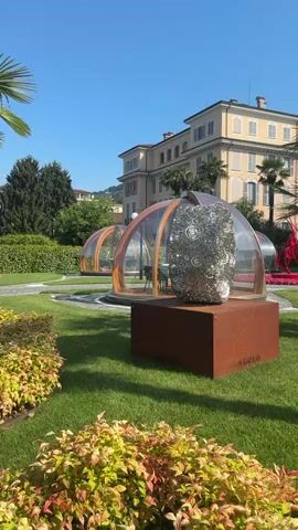 This media file captures a bright, sunny day in Binda, Italy. The scene is a well-manicured garden with lush green grass and vibrant foliage. In the foreground, a low hedge with red and yellow leaves lines the bottom of the frame.

The primary subjects are several contemporary glass and wood geodesic domes, appearing to be outdoor dining pods, scattered across the lawn. One dome in the foreground contains a large, metallic, abstract sculpture with a spiraling, intricate design. Another dome further back is furnished with a table and chairs, suggesting its use for dining. A bright red abstract sculpture is visible behind the domes, adding a splash of color to the landscape.

Palm trees and other lush vegetation are interspersed throughout the garden. In the background, a large, elegant building with a pale yellow facade and multiple windows stands under a clear blue sky. The architecture suggests a luxurious villa or hotel.

The time of day is clearly midday, given the bright sunlight and the long shadows cast by the structures and palm trees. The weather is fair and pleasant, ideal for outdoor activities. There are no people visible in the media file. The overall mood is serene and sophisticated, highlighting a blend of art, architecture, and nature.

Visible text includes the word "AKELO" inscribed on the base of the foreground dome's pedestal.