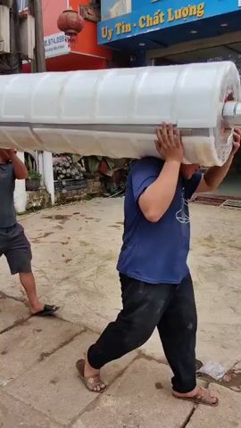 Three men are carrying a long, white, roughly cylindrical object on their shoulders. They are moving down a paved street in what appears to be a commercial or residential area. The object is quite large, almost appearing like a section of a pipe or some sort of prefabricated building component, wrapped in plastic. 

The men are dressed casually in t-shirts, shorts, and sandals or flip-flops. The environment consists of buildings, parked cars (a red one is visible), and signs in Vietnamese. The object is being moved towards the back of a small blue pick-up truck labeled "Porter 150" with the brand name "Hyundai".
