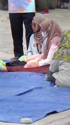 The video shows a group of people sitting on a blue tarp on the ground outdoors. In the foreground, a person's legs are visible, with blue and green covers on their feet. They are lying on a brown cloth. Two young women wearing hijabs are seated next to each other, facing the person on the ground. One woman is wearing sunglasses and a light-colored sweatshirt with "NEW" visible on it. The other woman is wearing a light pink shirt and a face mask. They are holding pink plastic bags. Another person, partially visible on the right, is wearing a green and brown patterned jacket and is also seated. In the background, another person stands wearing a black shirt and dark pants. The environment appears to be an outdoor, paved area with some foliage and what looks like a motorcycle to the left. The women are interacting with each other and the person on the ground, with one of them laughing.