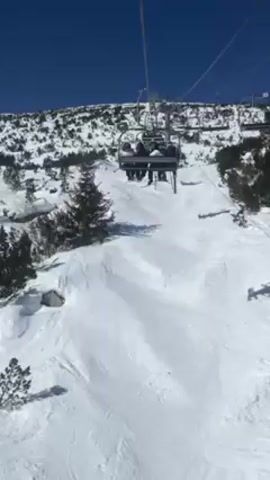 The video shows a view from a ski lift ascending a snow-covered mountain on a sunny day. We see other skiers on a lift car passing by. The landscape consists of snowy slopes interspersed with small pine trees and bushes. The sky is clear blue with some white clouds. The camera's perspective is from within one of the ski lift cars.