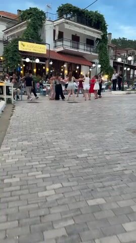 People are dancing in a circle in a cobblestone plaza in front of a restaurant called Alexandras Taverna Restaurant. The people are holding hands and moving in unison. There are tables and chairs set up outside the restaurant, and some people are sitting and watching the dancers. The sky is cloudy, and the lighting suggests it is either dusk or dawn. There are plants growing on the side of the building and string lights are visible. In the background, more buildings and trees are visible.