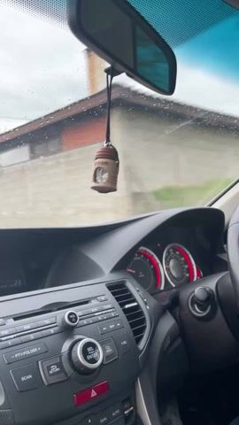 The video shows a car driving down a street in a village during light rain. The view is from the driver's seat, focusing on the road ahead through the windshield, which has raindrops on it. The car's dashboard and interior are visible, including the radio display showing a station and the steering wheel. The exterior shows houses with green fences and bare trees under a cloudy sky. The overall scene is of a rainy day drive in a rural or suburban setting.