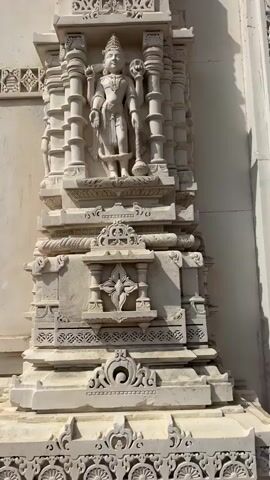 A close-up view shows intricate white marble carvings on the exterior of a temple. A statue of a male deity, possibly Vishnu or Krishna, stands in a niche supported by columns. The deity holds a mace or staff in one hand. The carving displays detailed ornamentation, including floral patterns and geometric designs. The camera pans upward, revealing the vast wall of the temple and a doorway with similar decorative carvings. Sunlight casts shadows on the wall, indicating it is daytime.