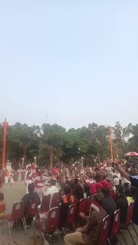 A crowd of people are seated on red chairs in the foreground, facing away from the camera. They appear to be watching a performance in a park. In the background, performers in red and white costumes are dancing. Trees and a tall orange structure are visible behind the performers. The sky is overcast.