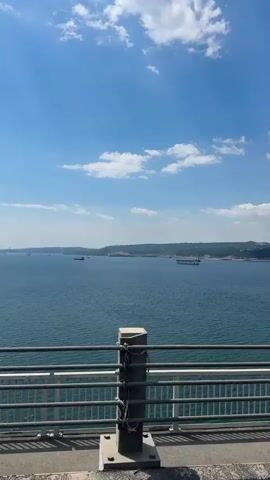From a fixed position at the edge of a bridge, the camera captures an open view of a vast expanse of water under a bright, blue sky dotted with scattered white clouds. In the distance, a few ships are visible on the water, along with a coastline featuring rocky land formations. The bridge's gray metal railing and supports are in the foreground, partially framing the expansive seascape. A dark pole stands upright near the bridge railing, momentarily obscuring the view as the camera pans slightly to the left, revealing a wider view of the ocean. There is a strong wind.