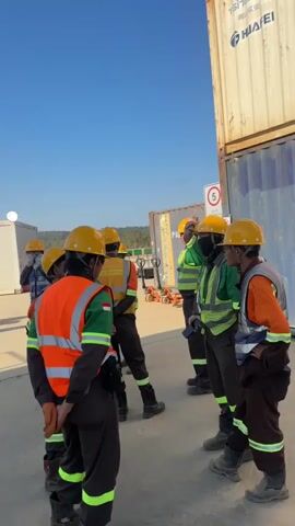 A group of construction workers in orange and green safety vests and yellow hard hats stand in a circle outdoors under a clear blue sky. Several of the workers are holding clipboards and appear to be in a briefing or discussion. In the background, there are modular buildings, shipping containers, and some construction equipment. The ground is paved and appears to be a work site.