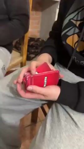 A person in a black sweatshirt and grey pants holds a deck of red cards. The cards have "MARTINI" printed in a white vertical bar on their backs. The person shuffles the deck. Another person is sitting to the left. The person with the cards then laughs.