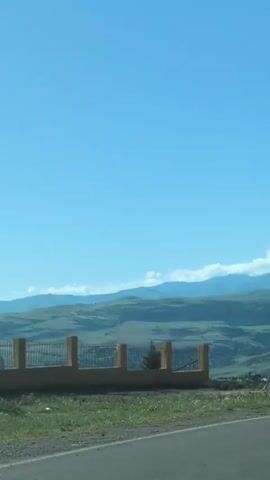 The video shows a scenic view from a car driving on a road. The road curves to the right, and there is a white barrier on the side. Beyond the road, there are rolling green hills covered with trees and shrubs. In the distance, there are mountains, some with snow-capped peaks. The sky is a clear blue with a few scattered clouds. The time stamps indicate the progression of the view as the car moves along. There are no people or animals visible in the video.