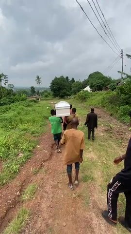 A group of men are carrying a white coffin on their shoulders up a dirt path. There are trees and vegetation on either side of the path, and a cloudy sky above. In the background, there are some buildings and a valley. The men are dressed in casual clothing.