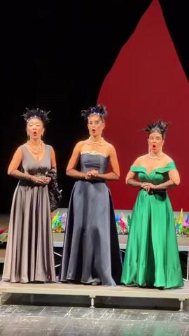 Three women stand on a stage, dressed in formal gowns and performing. The woman on the left wears a grey gown and opera gloves. The woman in the middle wears a dark blue strapless gown and opera gloves. The woman on the right wears a green off-the-shoulder gown and opera gloves. Behind them, a man dressed in a vibrant, colorful, feather-adorned costume stands and sings. The stage has a large red backdrop.