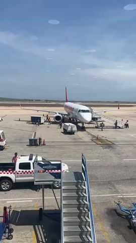 Here's a description of the video:

The video shows an Embraer 190 aircraft operated by Conviasa at an airport. Several ground personnel are around the aircraft, potentially preparing for departure or arrival. A set of airstairs is positioned near the front of the plane, and people are using it.  A truck labeled "Aeropostal" is parked nearby. The weather appears to be clear with some clouds.  The video is taken from an elevated position, likely inside the airport terminal.